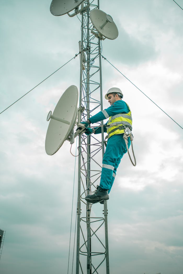 services-01 A worker in safety gear installs a satellite dish on a telecom tower under an overcast sky.